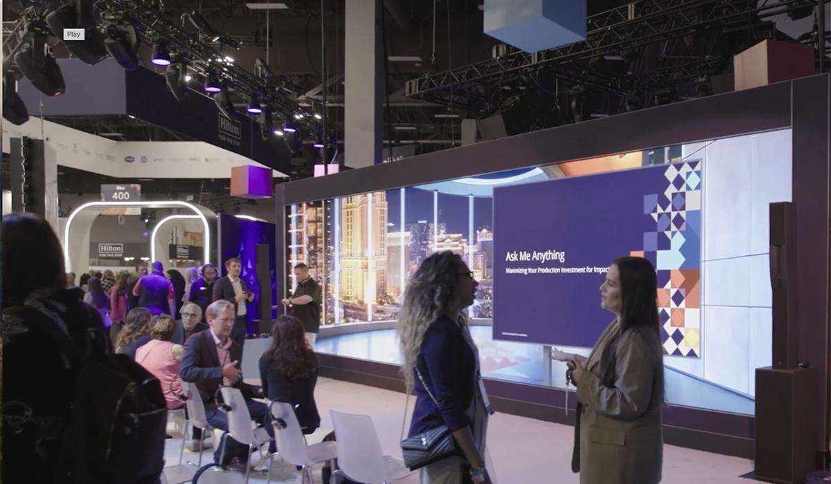 People gather in front of a large screen displaying Ask Me Anything at an indoor event or conference. Some attendees are seated while others stand and converse under bright lighting.