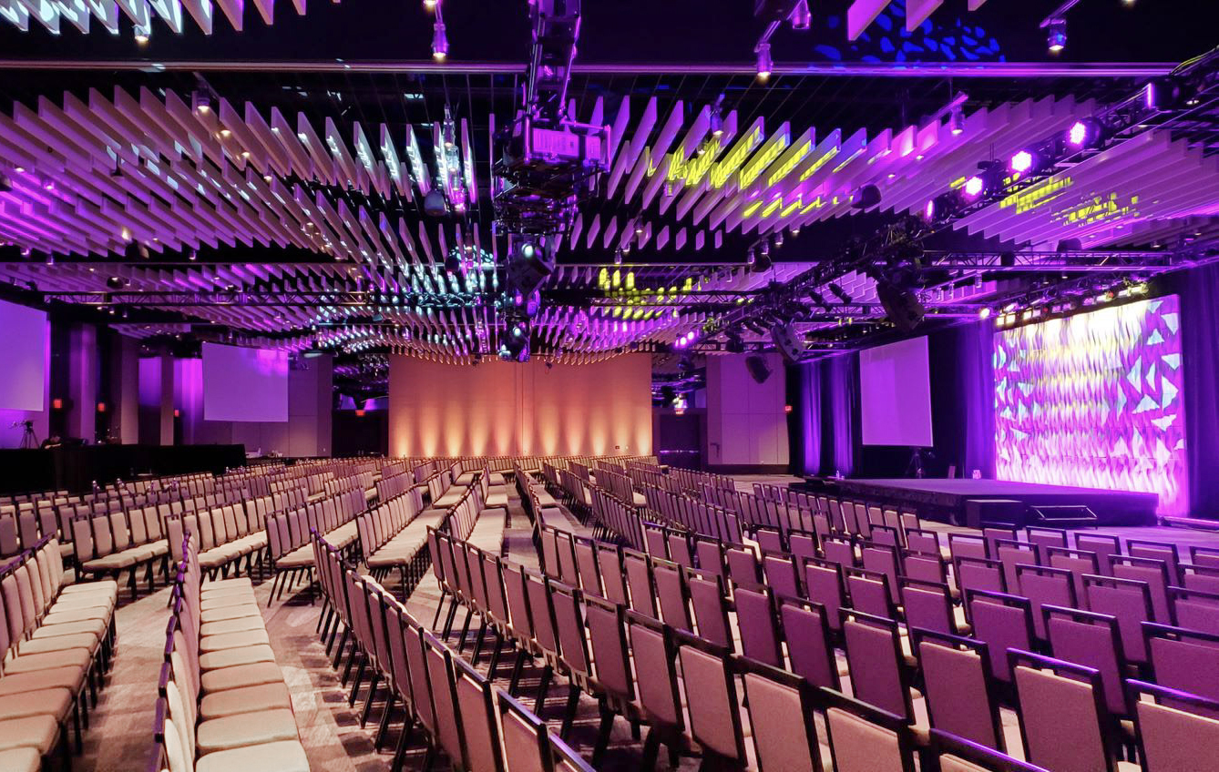 Rows of empty chairs are arranged in front of large, brightly lit stages under a ceiling with geometric panels and purple lighting, creating a modern and vibrant conference or event space.