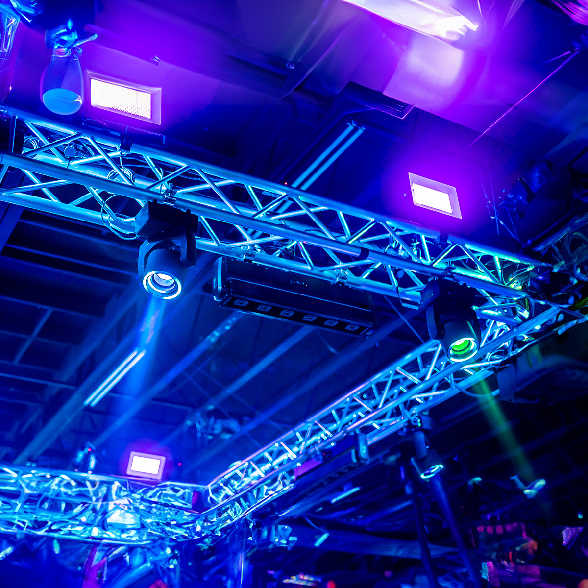 A close-up of a metal truss structure with mounted stage lights glowing in blue and purple hues, creating a vibrant, illuminated atmosphere, with ceiling pipes visible in the background.