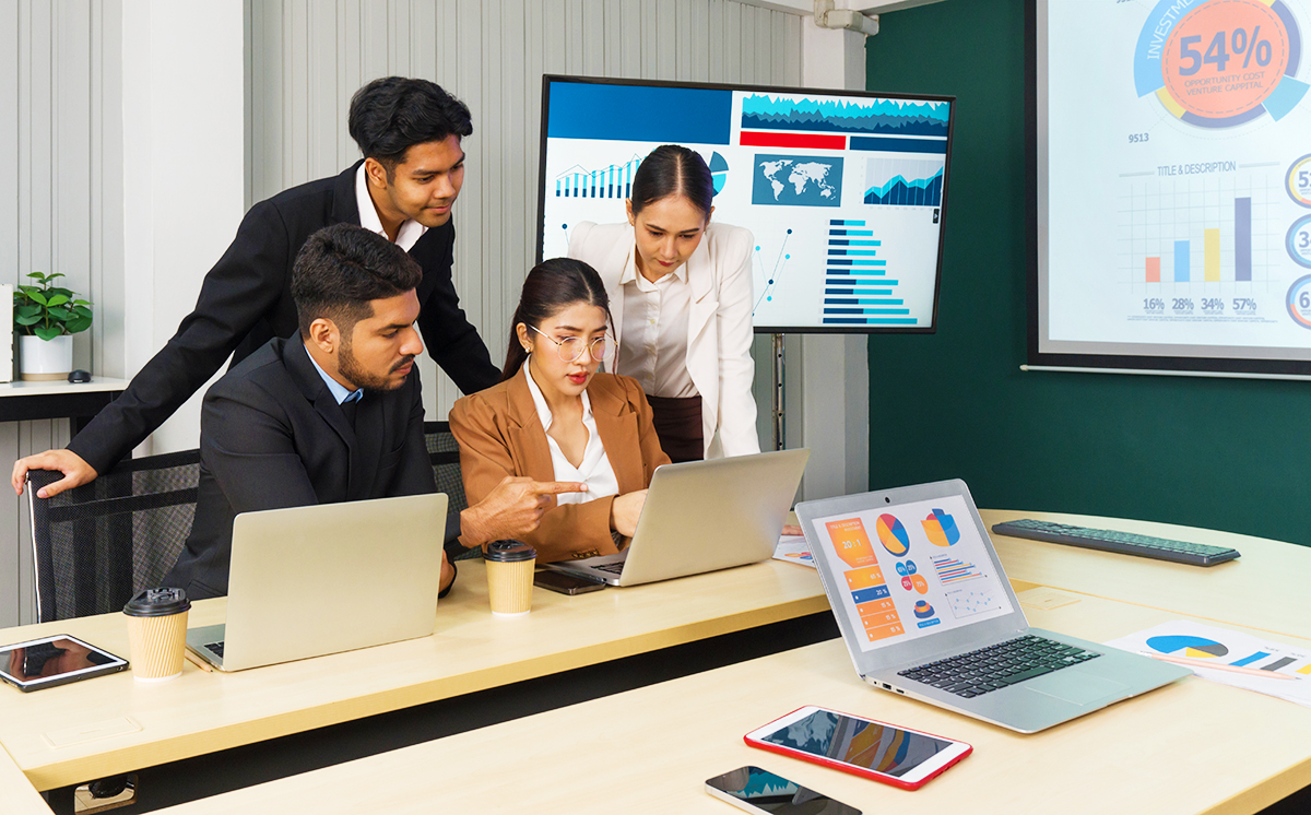 Four business professionals collaborate at a table with laptops, charts, and coffee cups, analyzing data on screens and printed reports in a modern office meeting room.