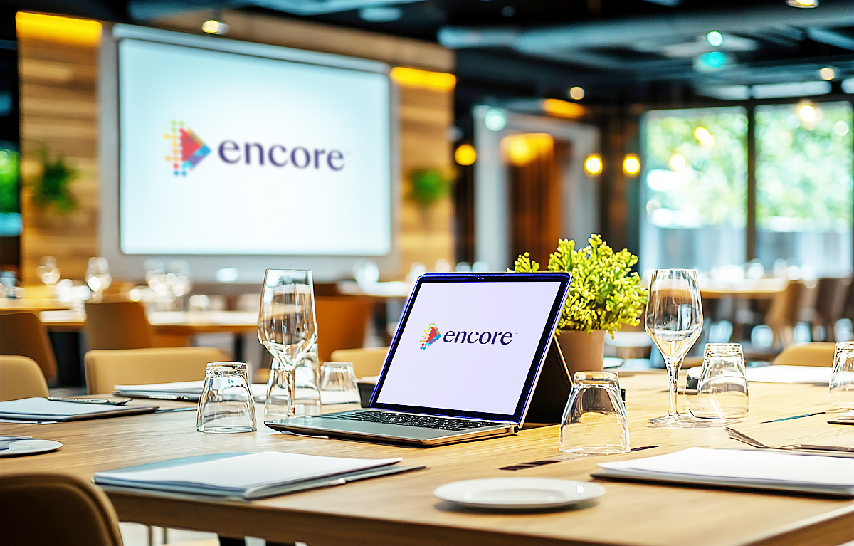 A modern conference room with a laptop and glassware on a table, all set for a meeting. The laptop and a large screen in the background display the Encore logo. The room is stylishly decorated with plants and wooden accents.