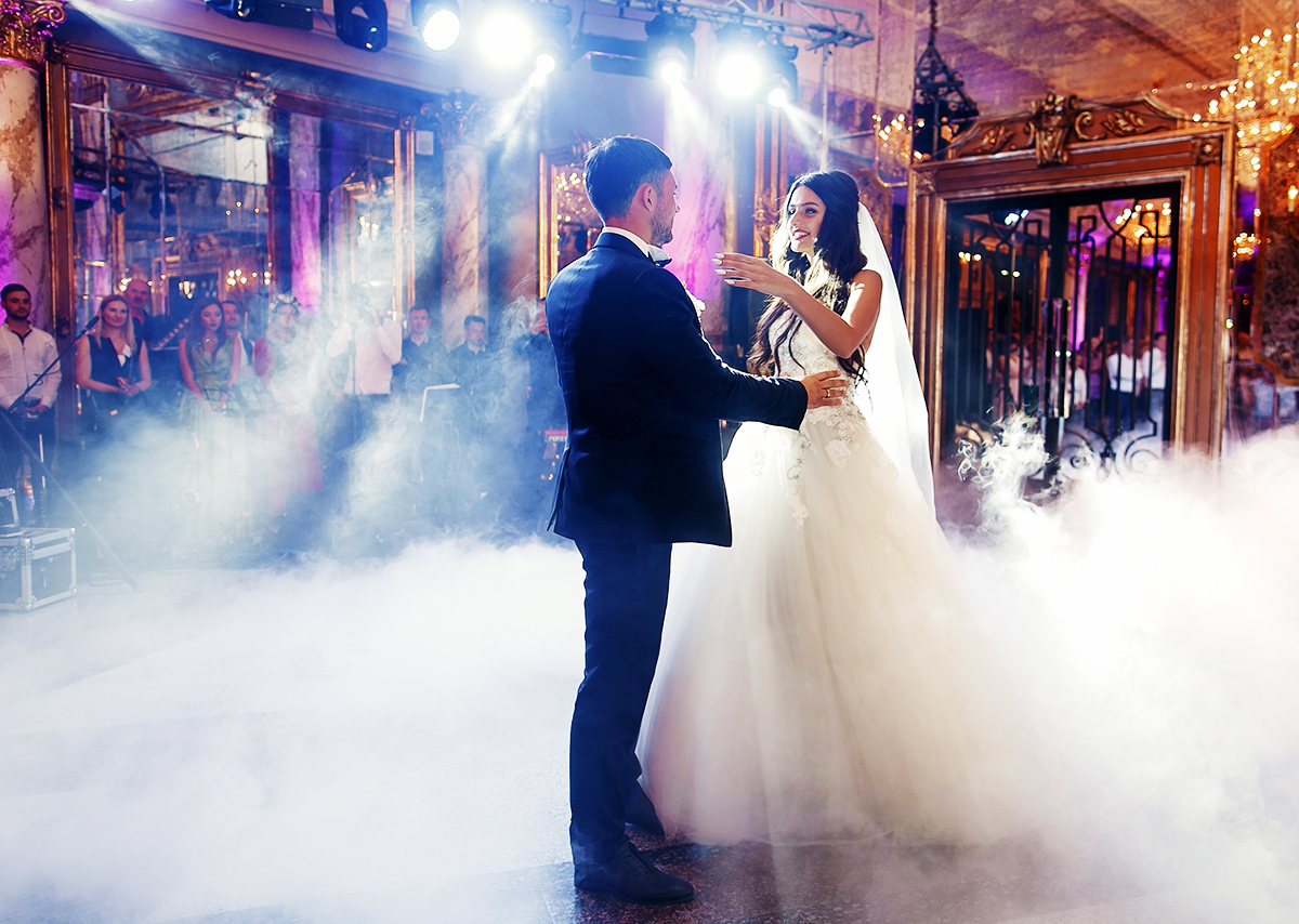 A bride and groom share their first dance on a smoke-covered dance floor in a grand, elegant venue with chandeliers and purple lighting, while guests watch in the background.