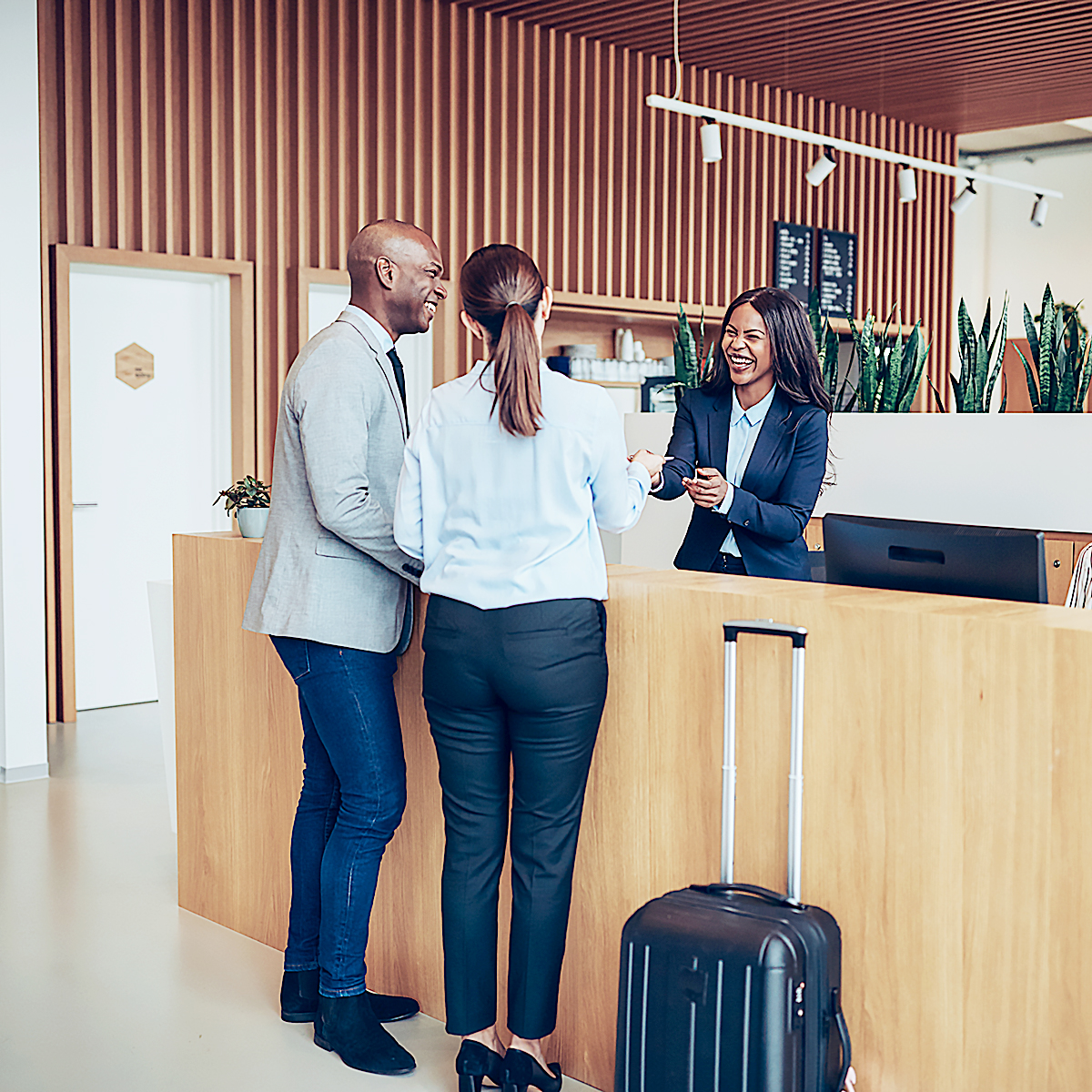 Two guests stand at a modern hotel reception desk, smiling and talking with a receptionist. A suitcase is next to them, and the lobby features wooden accents and green plants.