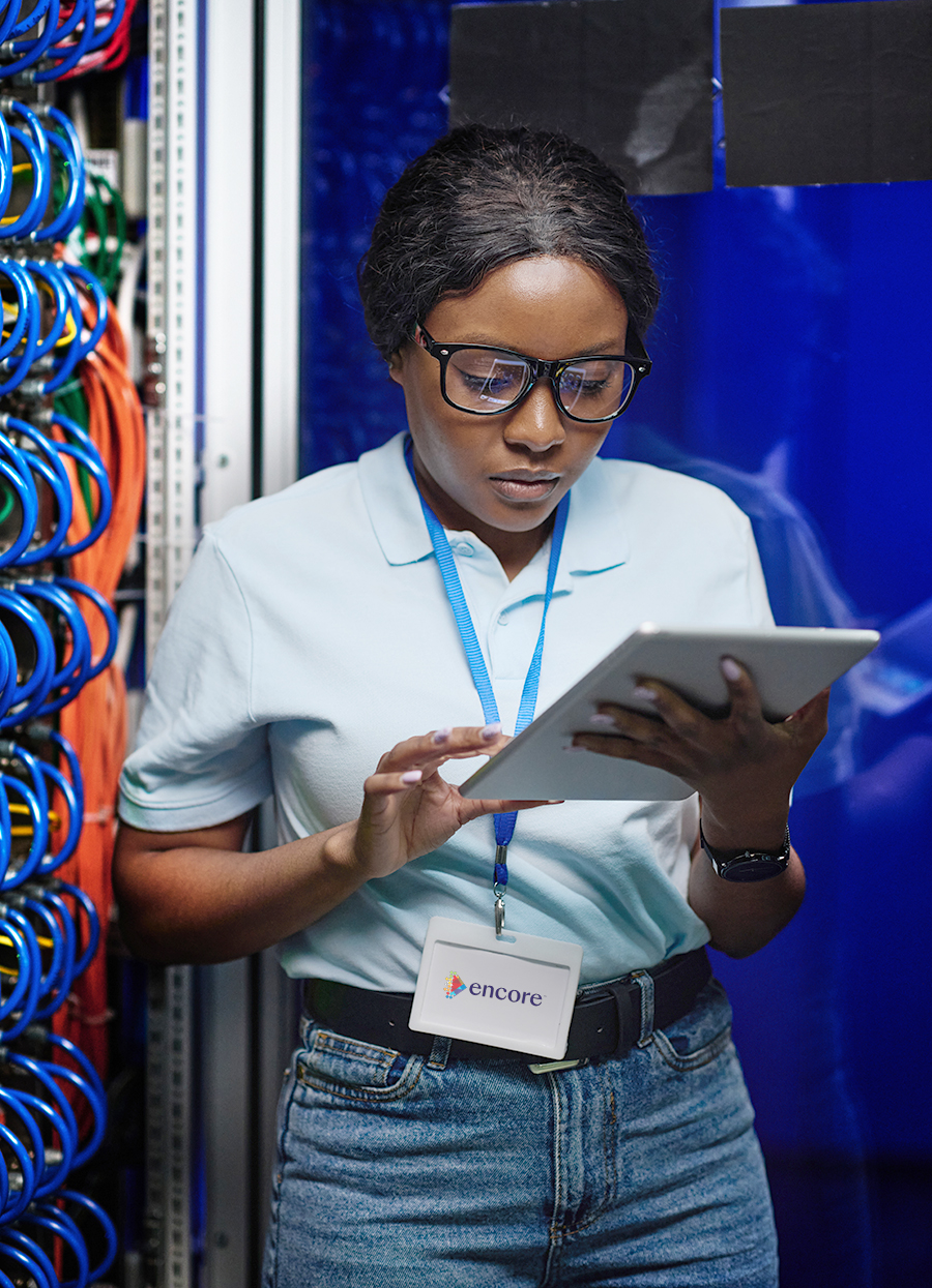 A person wearing glasses and a name badge reading encore stands in a server room, using a tablet. Colorful network cables are visible in the background. The person appears focused on the device.