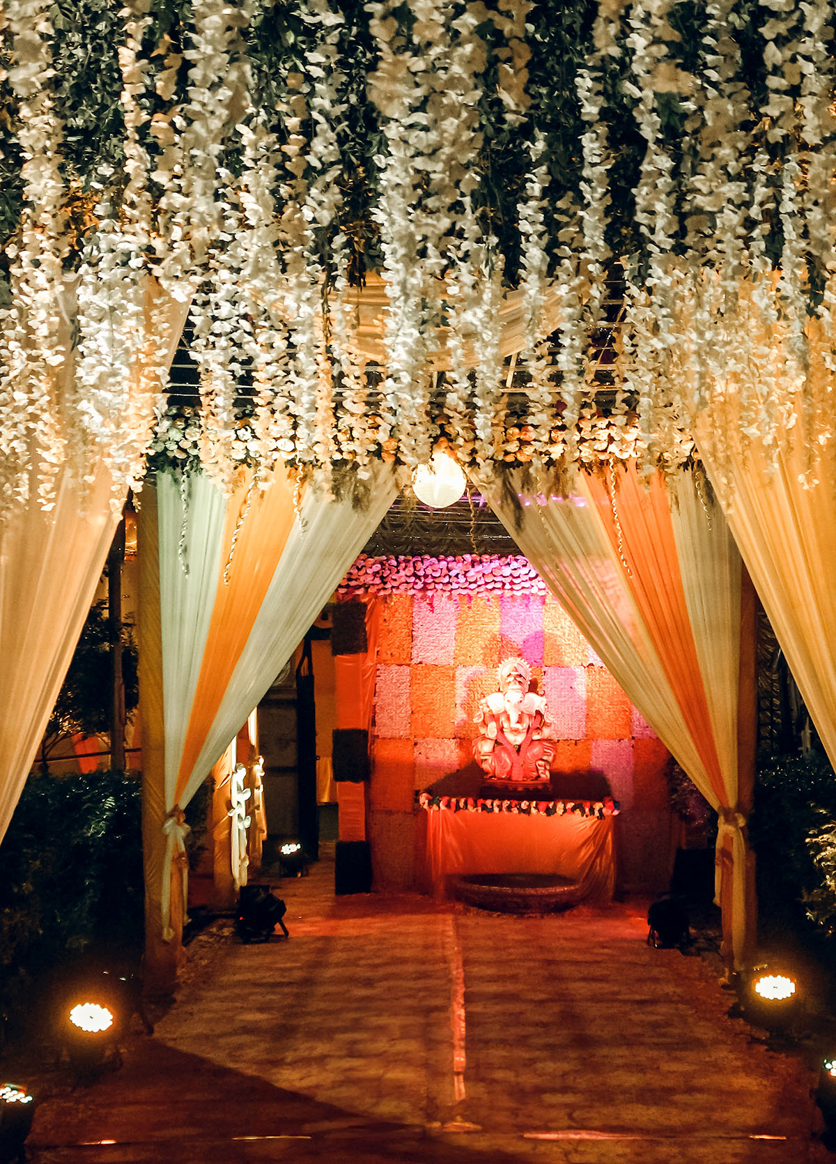 A decorated walkway with draped white fabric and hanging flowers leads to an illuminated stage featuring an idol of the Hindu deity Ganesha, surrounded by warm lights and vibrant decor.