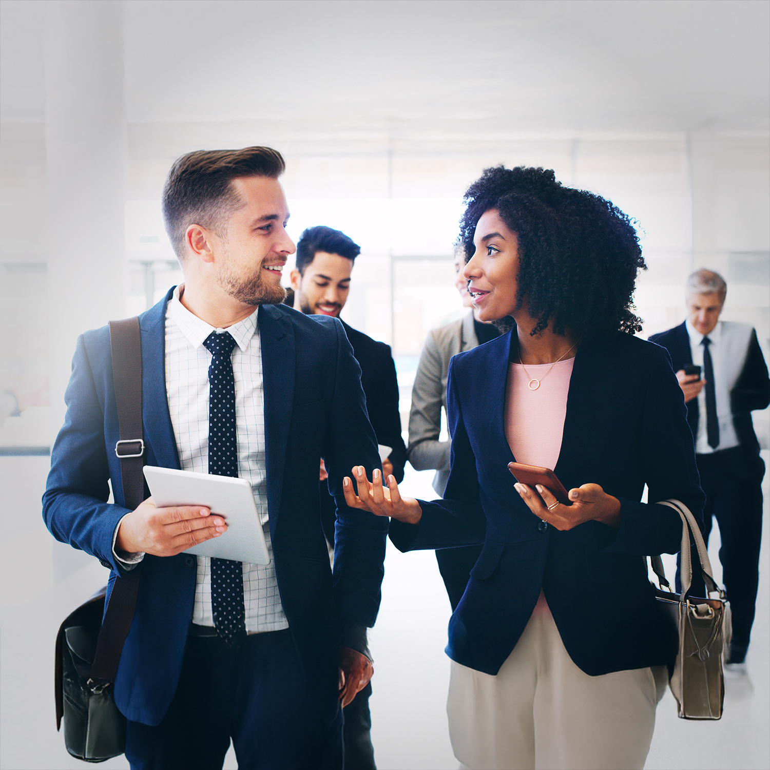 Two business professionals walk and chat in an office hallway, holding a tablet and phone. Other colleagues walk behind them, all dressed in business attire. The atmosphere appears collaborative and professional.