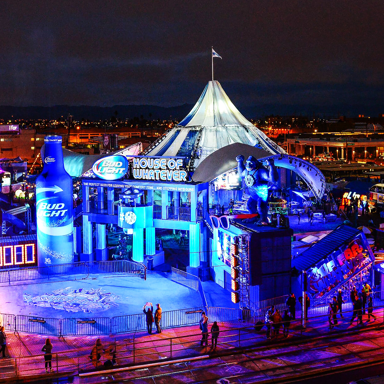 A brightly lit event venue at night features a large tent with House of Whatever signage, glowing blue lights, a giant Bud Light bottle, and a gorilla statue, with people walking nearby.