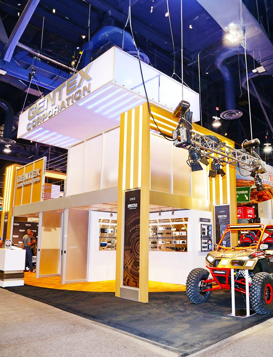 A brightly lit trade show booth for Gentex Corporation with yellow and white striped panels, tech displays inside, and a red off-road vehicle in front. Overhead, a camera rig is suspended from the ceiling.