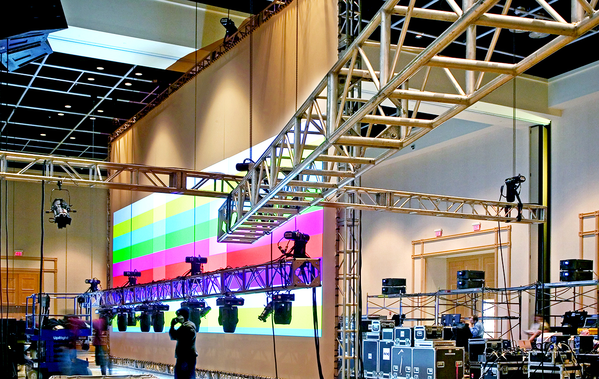 A large event space with stage lighting trusses, spotlights, and audiovisual equipment set up in front of a wall displaying a colorful test pattern screen, preparing for a production or event.