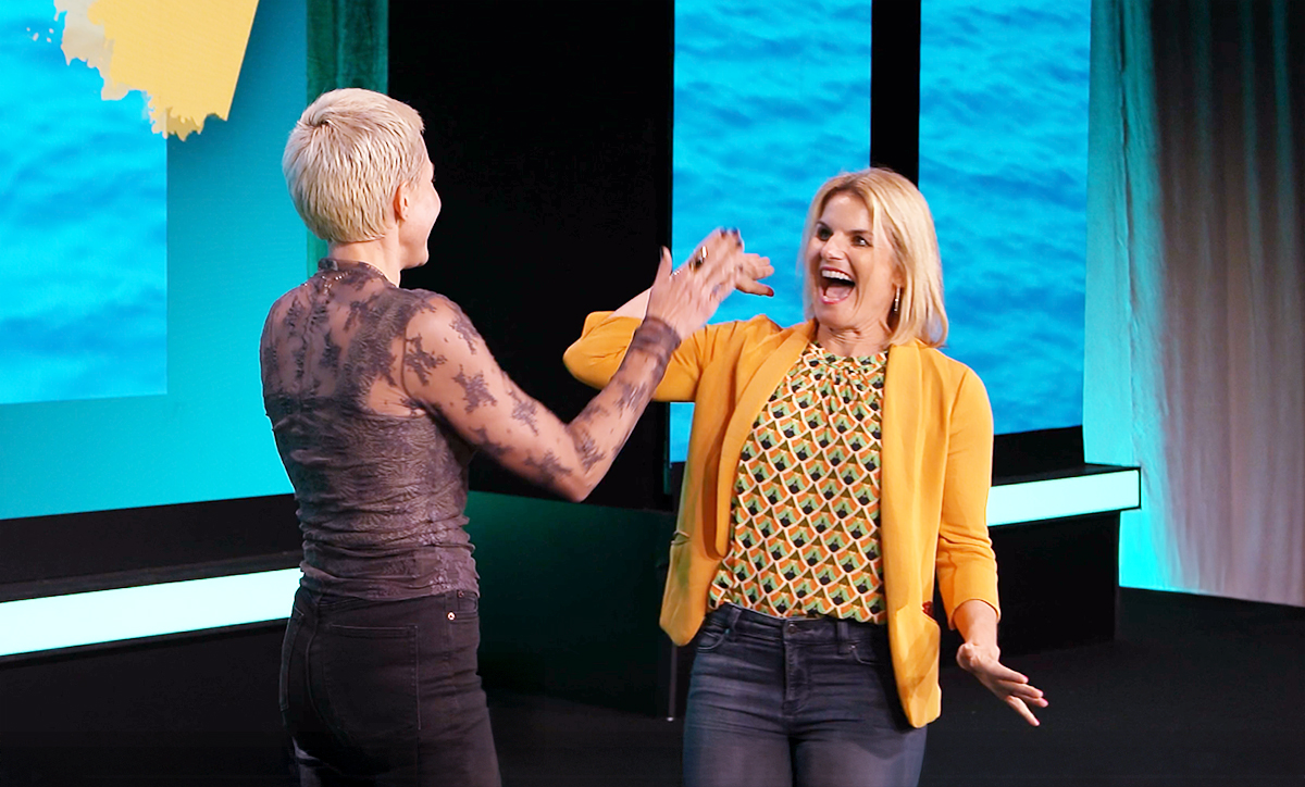 Two women smiling and giving each other a high five on a brightly lit stage with turquoise and yellow accents in the background. One woman wears a yellow blazer, the other a sheer black top.