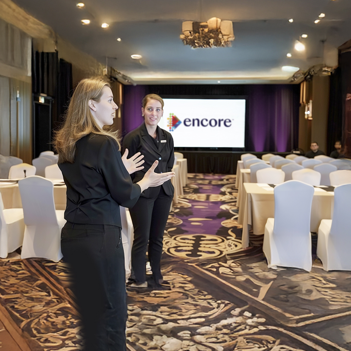 Two women in black uniforms stand and talk in a conference room with white chair covers. A large screen at the front displays the Encore logo. The room is set up for a presentation or event.