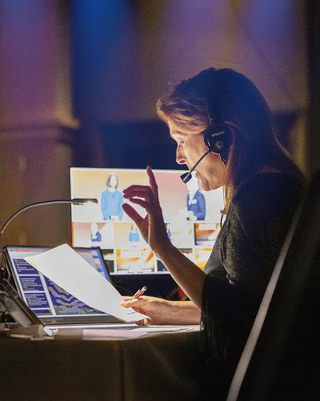 A woman wearing a headset sits at a desk with a laptop and papers, speaking and gesturing while looking at multiple monitors showing people on video calls.